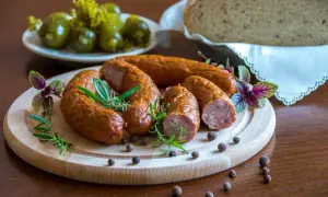 Kiełbasa podwawelska na desce z ziołami, obok ogórki i chleb.