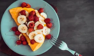 Omlet z bananem i malinami, polany syropem, na jasnoniebieskim talerzu. Obok leży widelec.