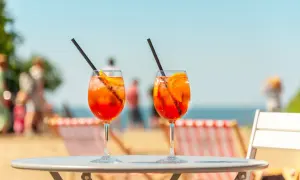 Dwa drinki Aperol Spritz z plastrami pomarańczy i słomkami na stole, w tle plaża i morze.