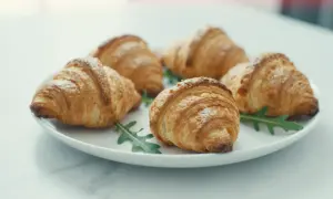 Pięć puszystych croissantów z ciasta francuskiego, posypanych cukrem pudrem, ułożonych na talerzu z rukolą.