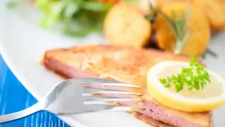 Chrupiąca mortadela w panierce, podana z ziemniakami i sałatką, ozdobiona plasterkiem cytryny i natką pietruszki.