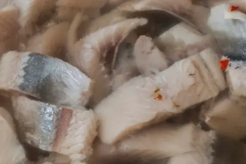 Kawałki kiszonych śledzi w zalewie, z widocznymi fragmentami skórki i przyprawami.