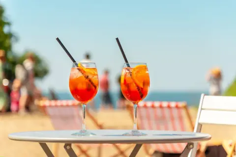 Dwa drinki Aperol Spritz z plastrami pomarańczy i słomkami na stole, w tle plaża i morze.