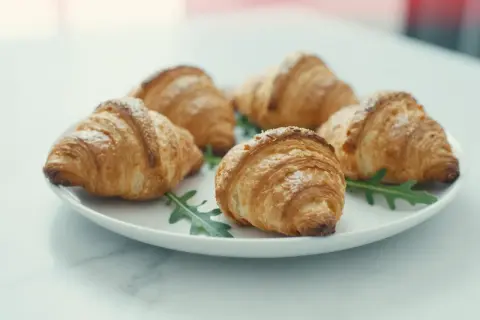 Pięć puszystych croissantów z ciasta francuskiego, posypanych cukrem pudrem, ułożonych na talerzu z rukolą.