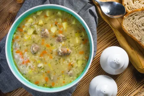 Aromaticzny krupnik, przepis babci, z kawałkami mięsa, ziemniaków i marchewki, posypany koperkiem. Obok kromki chleba i solniczki.