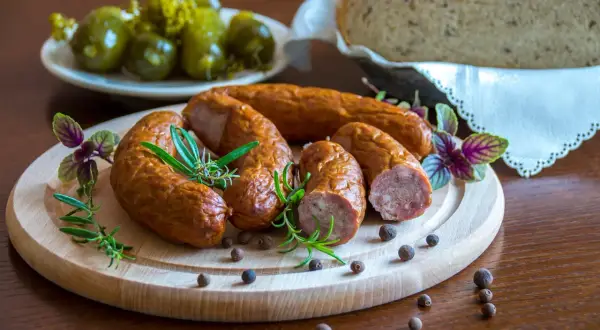 Kiełbasa podwawelska na desce z ziołami, obok ogórki i chleb.