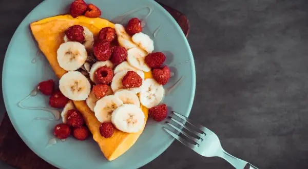 Omlet z bananem i malinami, polany syropem, na jasnoniebieskim talerzu. Obok leży widelec.