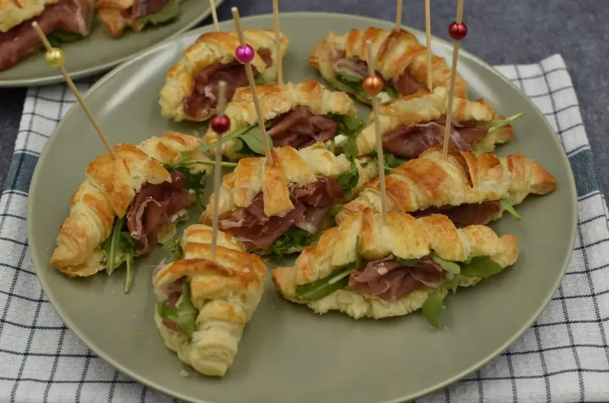 Mini croissantów z ciasta francuskiego z szynką parmeńską i rukolą, udekorowane kolorowymi patyczkami.