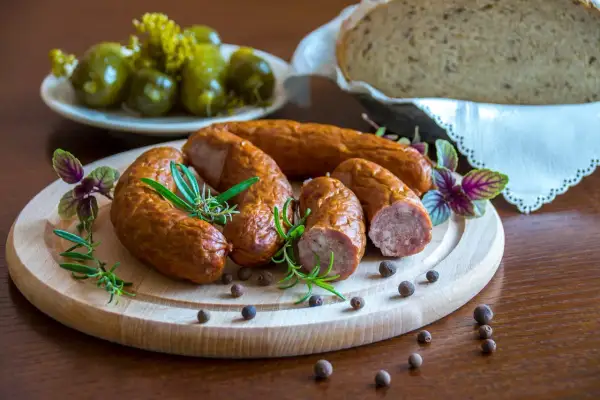 Kiełbasa podwawelska na desce z ziołami, obok ogórki i chleb.