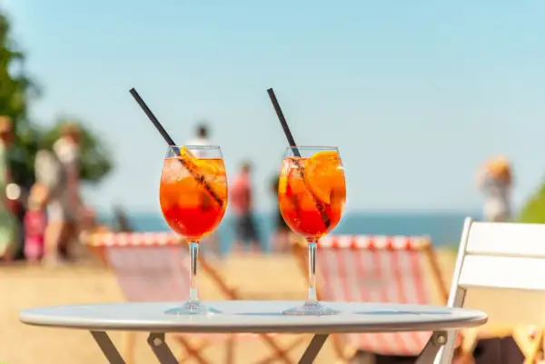 Dwa drinki Aperol Spritz z plastrami pomarańczy i słomkami na stole, w tle plaża i morze.