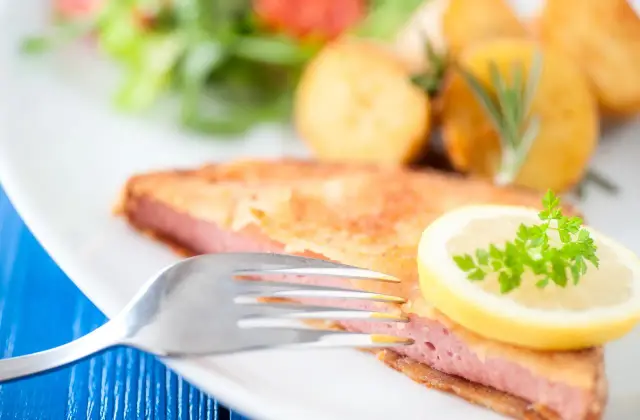 Chrupiąca mortadela w panierce, podana z ziemniakami i sałatką, ozdobiona plasterkiem cytryny i natką pietruszki.